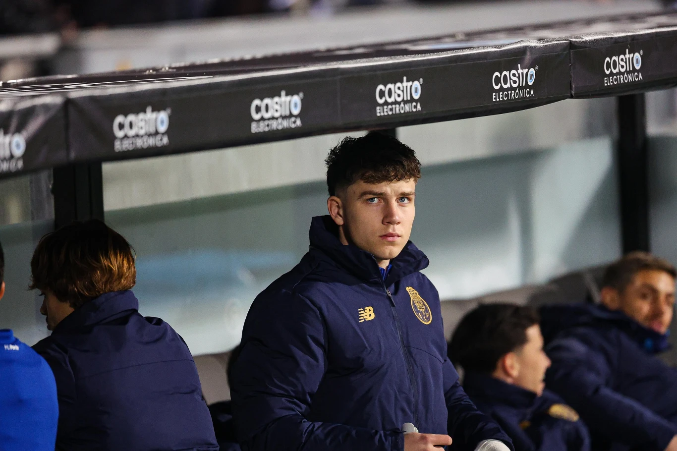 Młody mężczyzna w kurtce sportowej siedzi na ławce rezerwowych podczas wydarzenia sportowego, wokół niego znajdują się inni zawodnicy w podobnych strojach, a nad nimi widoczny jest daszek z reklamami.