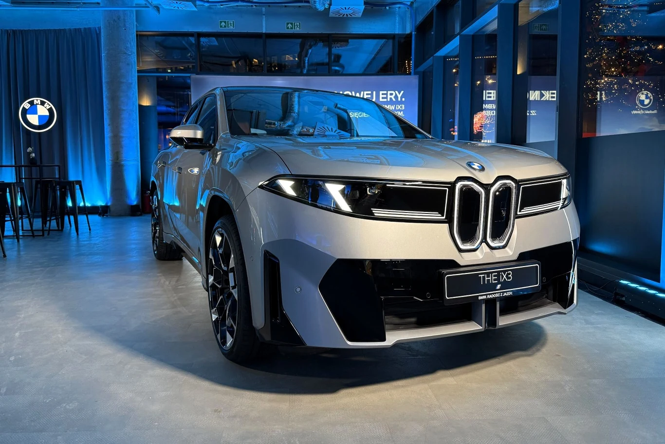 Nowe BMW iX3 to pierwszy model marki stworzony w ramach koncepcji Neue Klasse. Srebrny samochód BMW, model THE iX3, stoi oświetlony w nowoczesnym salonie z niebieskim oświetleniem ambientowym, w tle widoczne logo marki oraz industrialne wnętrze z dużymi oknami.