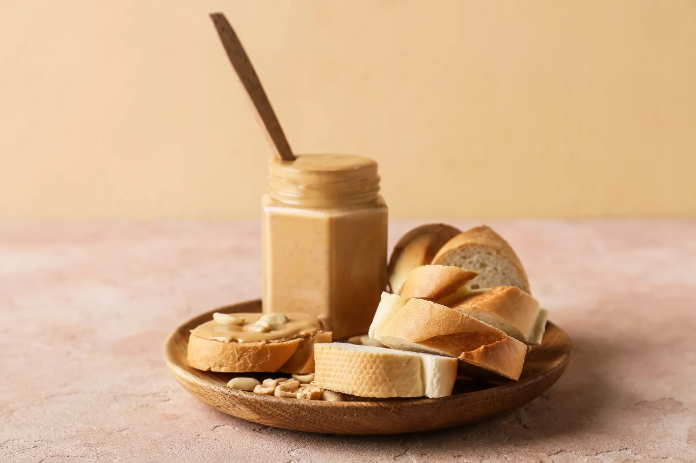 Szklany słoik z masłem orzechowym, drewniana łyżka wbita w masło, na drewnianym talerzu leżą podłużne kromki białego pieczywa, jedna z kromek posmarowana masłem orzechowym, wokół rozsypane orzeszki ziemne, pastelowe tło utrzymane w jasnych odcieniach.