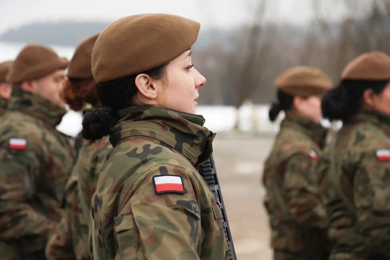 Program Ochrony Ludności i Obrony Cywilnej. Jakie działania realizuje rząd? Zdj. ilustracyjne Grupa żołnierzy w mundurach i beretach stoi na zewnątrz w szeregu, o wyglądzie zimowym, widoczna polska flaga na rękawach. Na pierwszym planie kobieta zwrócona bokiem, w tle lekko zamglony pejzaż.