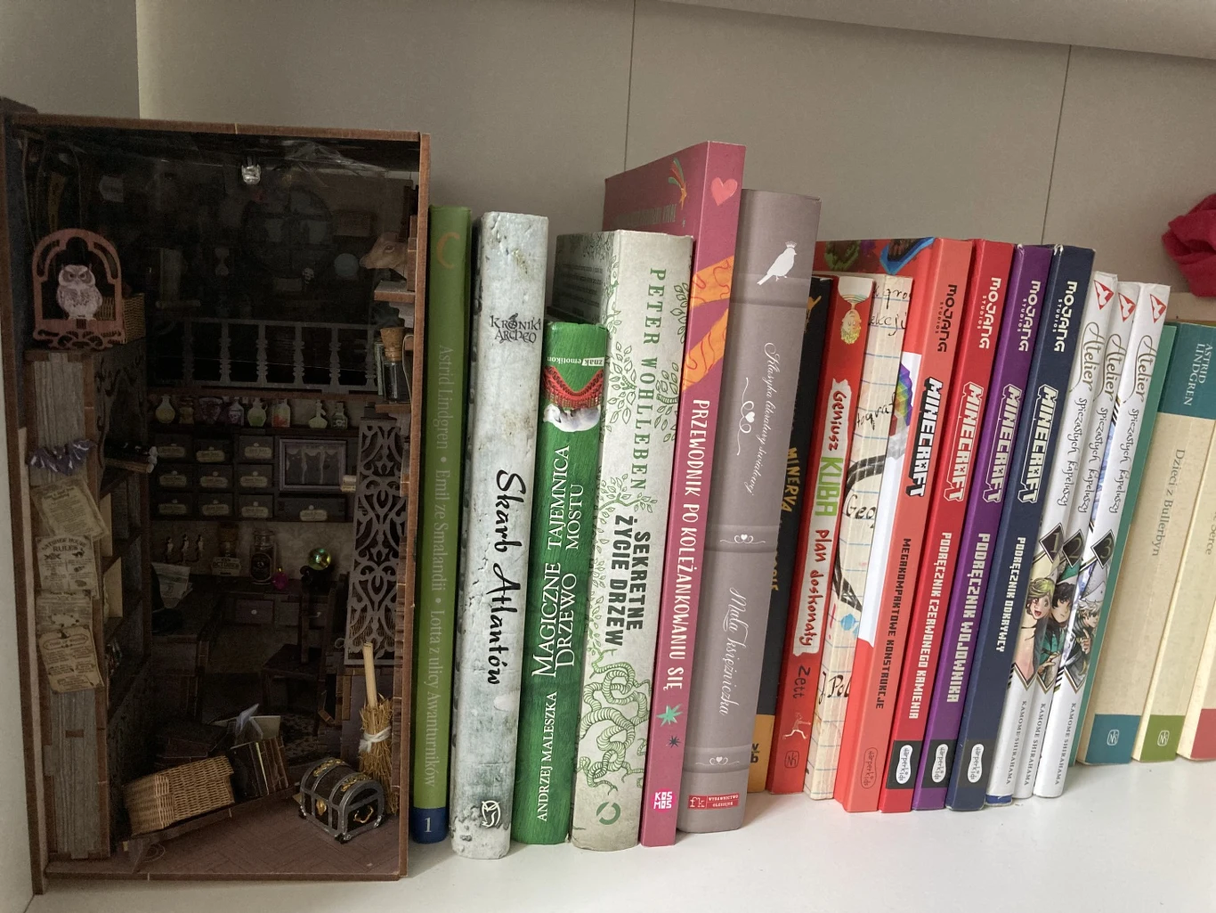 Rząd kolorowych książek ustawionych na białej półce, po lewej stronie znajduje się miniaturowa scena przypominająca wnętrze antykwariatu lub warsztatu, wykonana z papieru lub innych materiałów modelarskich.