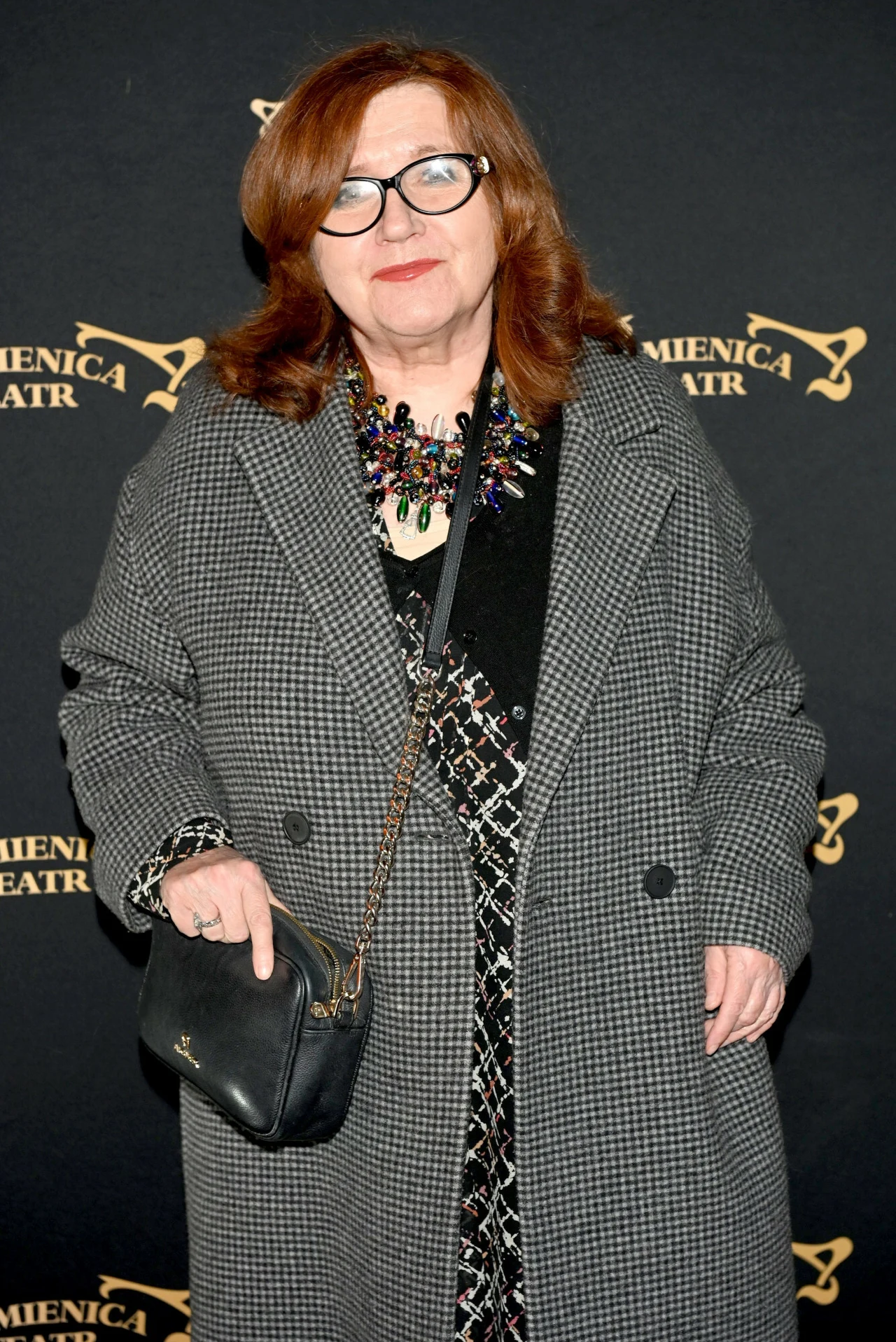 Ewa Ziętek Kobieta z rudymi włosami ubrana w elegancki płaszcz w kratę i ciemną sukienkę, z okularami na nosie oraz kolorowym naszyjnikiem, trzyma czarną torebkę na łańcuszku. Tło z logotypami teatru Kamienica.