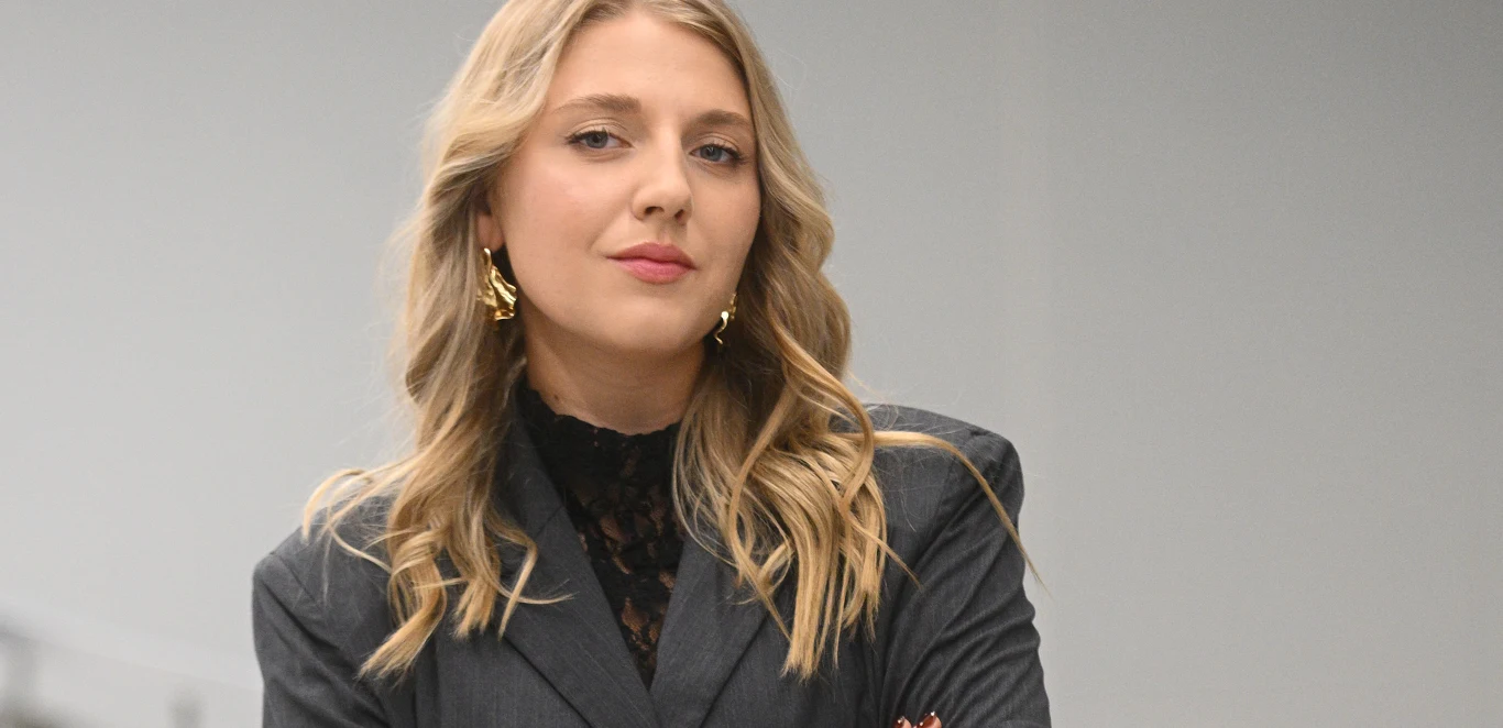 Maja Golec Kobieta o blond włosach w eleganckim, ciemnym garniturze z koronkową bluzką i złotymi kolczykami stoi na tle jasnego, neutralnego tła, prezentując pewność siebie i profesjonalizm.