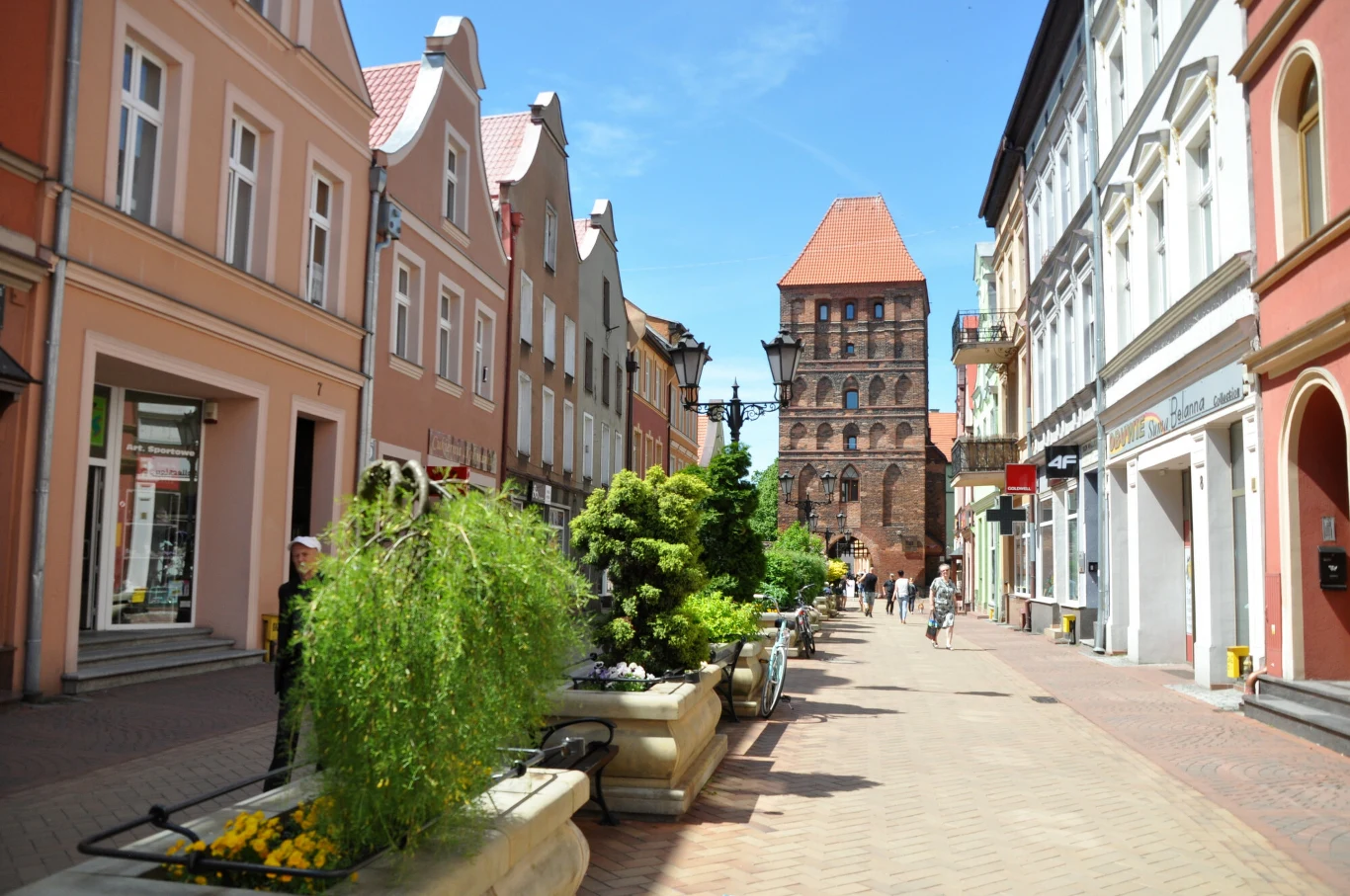 Ulica miasta z kolorowymi kamienicami i licznymi sklepami, piesi spacerują wzdłuż zadbanych donic z zielenią, w tle widoczna zabytkowa ceglana brama.