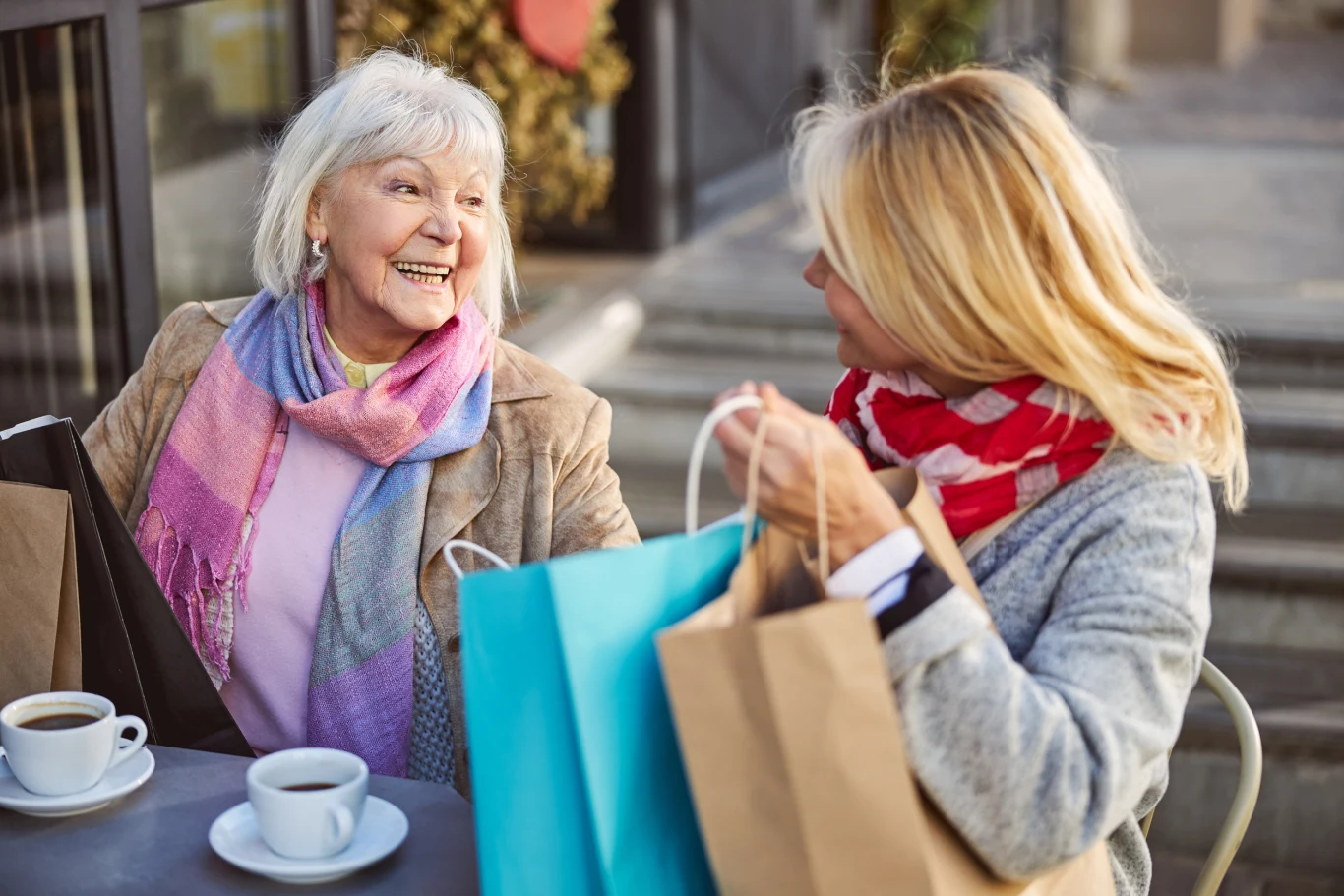 Dwie kobiety siedzą przy stoliku na zewnątrz kawiarni, rozmawiając i uśmiechając się do siebie. Przed nimi leżą torby z zakupami i filiżanki kawy. Kobiety są ubrane w ciepłe szaliki.