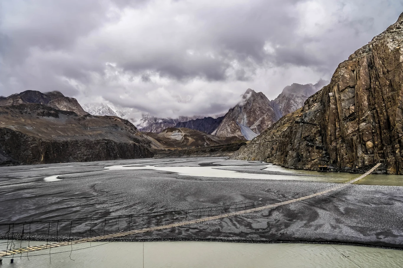 Pakistański most uznaje się za najbardziej niebezpieczny na świecie. Deski trzeszczą, a konstrukcja buja się na wietrze Wiszący most linowy rozciągający się nad szarą, kamienistą doliną, ciek wodny pod nim, otoczony surowymi, wysokimi górskimi szczytami częściowo przysłoniętymi chmurami.
