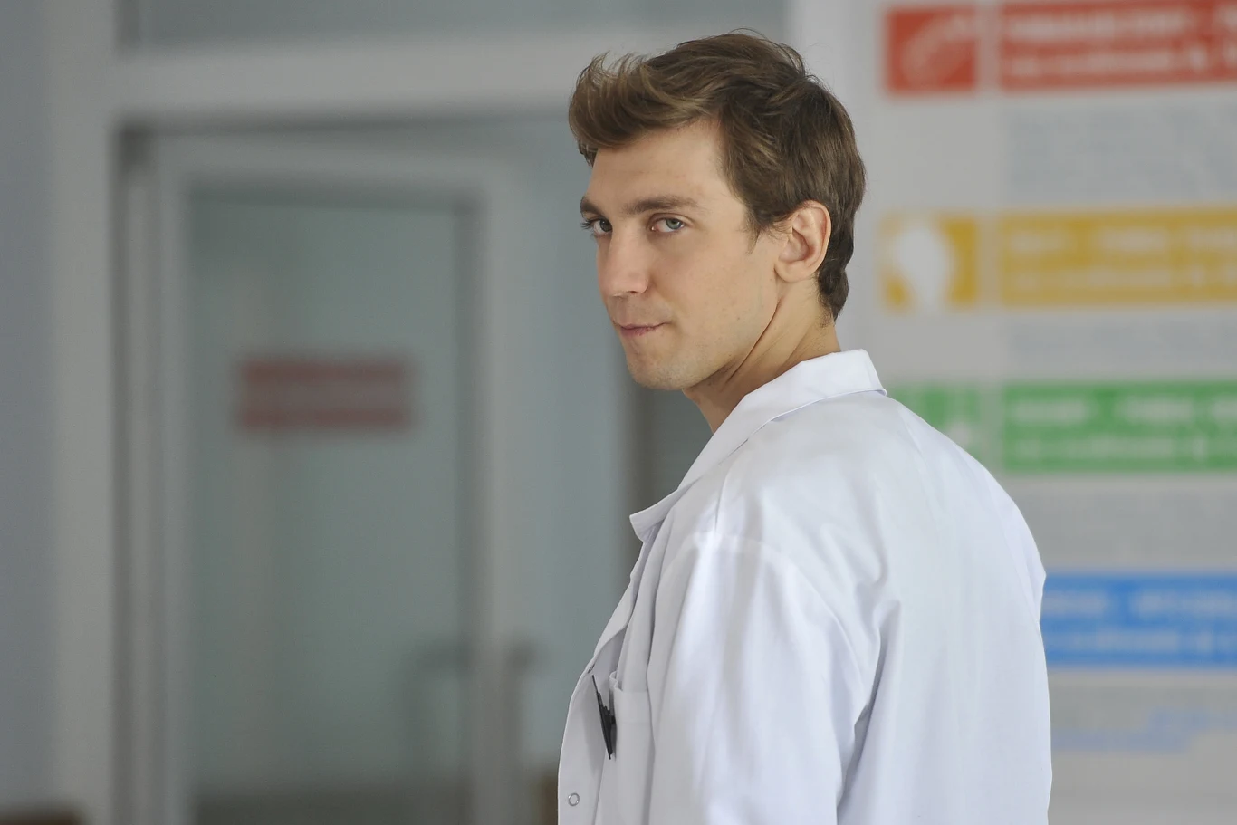 Młody mężczyzna w białym fartuchu medycznym odwraca się lekko, spoglądając na bok, tło częściowo rozmyte z kolorowymi planszami informacyjnymi na ścianie.