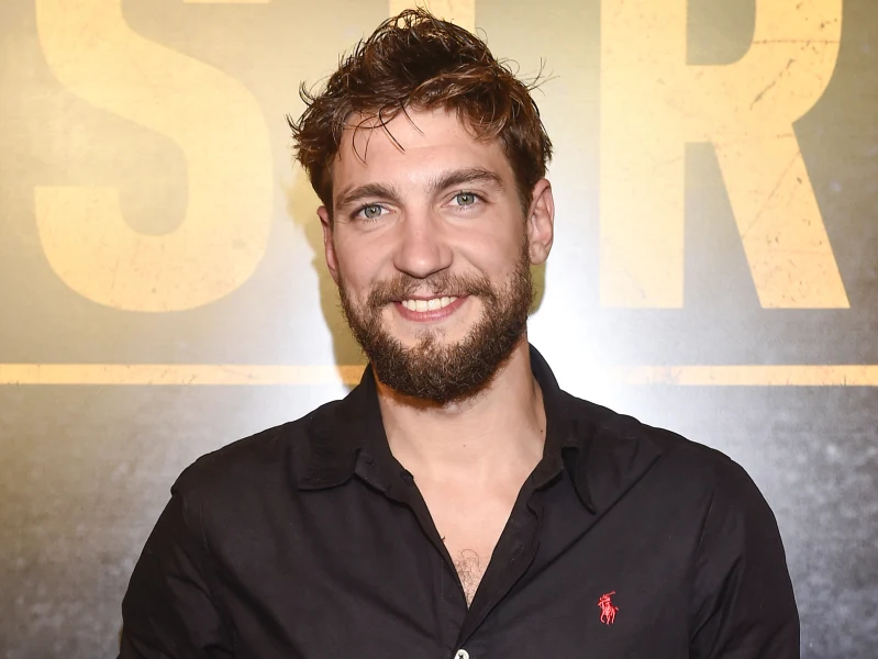 Mężczyzna z brodą i krótkimi, rozczochranymi włosami uśmiecha się, ubrany w czarną koszulę z emblematem marki Polo Ralph Lauren, znajduje się na tle grafiki z dużymi żółtymi literami.