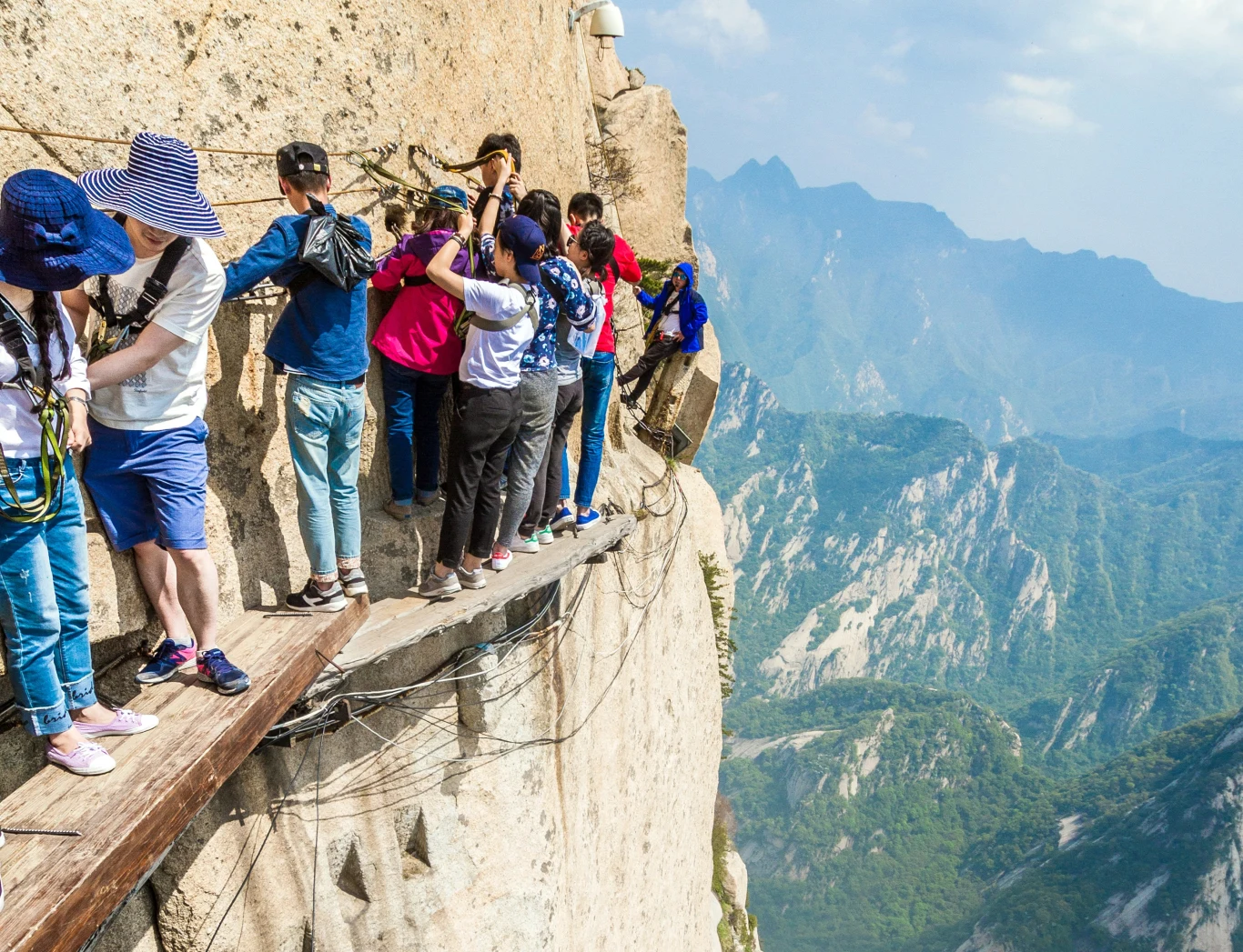 Turyści z całego świata ryzykują życiem, by wejść na "Świętą Górę". Co roku ginie tu mnóstwo osób Grupa turystów porusza się wzdłuż wąskiej, drewnianej kładki przymocowanej do stromego zbocza skalistej góry, trzymając się metalowych łańcuchów ochronnych; w tle rozciąga się widok na górzysty krajobraz.