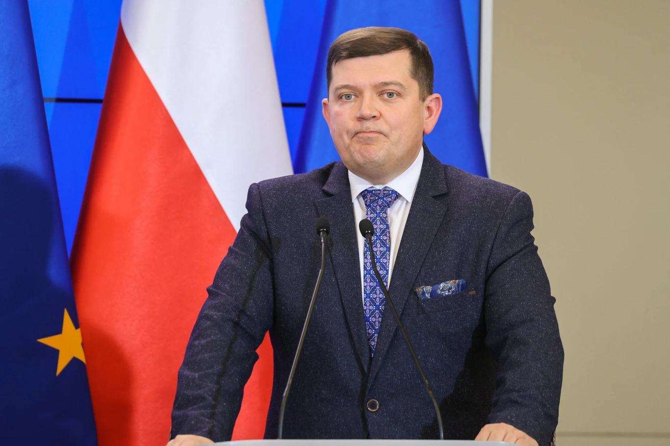 Mężczyzna w garniturze stojący za mównicą z dwoma mikrofonami, w tle widoczne flagi Polski i Unii Europejskiej, scena sugeruje wystąpienie publiczne lub konferencję prasową.