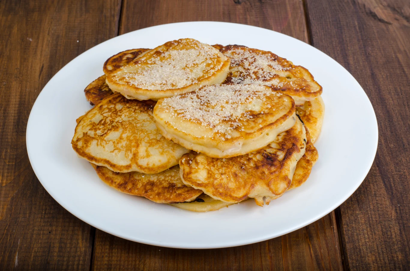 Stos puszystych placuszków na białym talerzu, niektóre posypane cukrem, ustawione na drewnianym blacie.