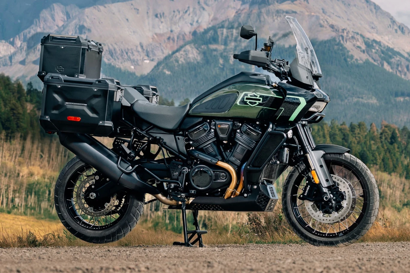 Motocykl typu adventure z dodatkowymi kuframi bocznymi i górnym, stojący na polnej drodze w górskim krajobrazie. W tle widać las i skaliste szczyty, a motocykl wyróżnia się nowoczesnym designem i solidnym wyposażeniem.