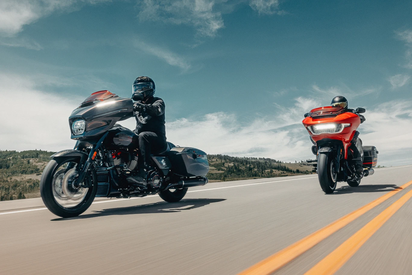 Dwóch motocyklistów w kaskach jedzie nowoczesnymi motocyklami szosowymi po asfaltowej drodze w otoczeniu natury. Szeroka jezdnia i błękitne niebo z lekkimi chmurami podkreślają swobodę podróży.