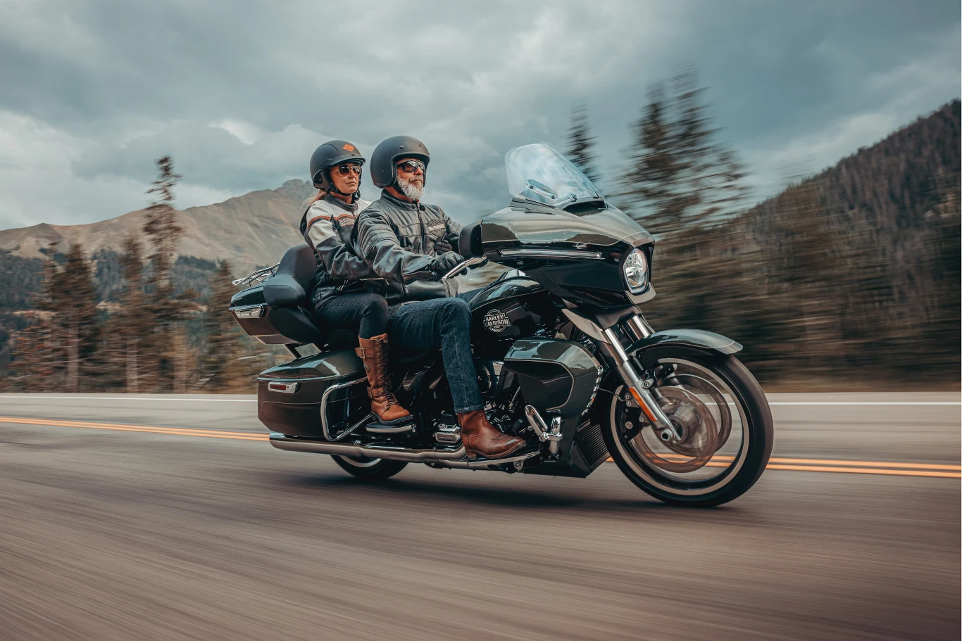 Dwie osoby w kaskach motocyklowych jadą motocyklem szosowym po asfaltowej drodze, otoczone górami i drzewami, ubrani w kurtki i spodnie motocyklowe.