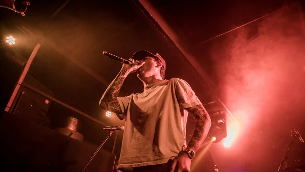 Wokalista na scenie trzyma mikrofon, ubrany w jasny t-shirt i czapkę z daszkiem, oświetlony intensywnym, czerwonym światłem. Tło wypełnione dymem scenicznym i sprzętem muzycznym, atmosfera koncertu klubowego.