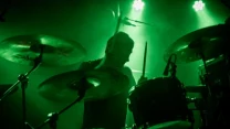 Perkusista grający na scenie, oświetlony zielonym światłem scenicznym, otoczony zestawem perkusyjnym i mikrofonem, w dynamicznej atmosferze koncertu.