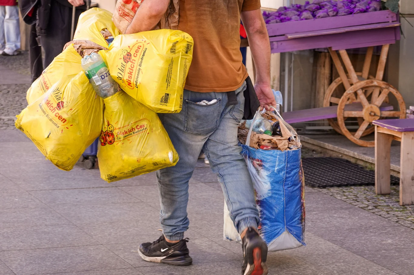 Mężczyzna niosący duże worki i torby wypełnione różnymi rzeczami, idący chodnikiem w pobliżu straganu z warzywami.