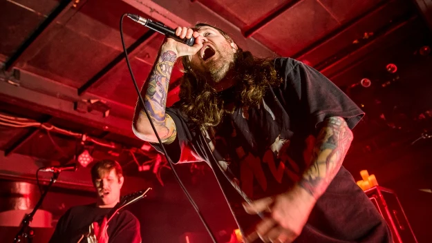 Wokalista z długimi włosami i tatuażami śpiewa do mikrofonu na scenie, w tle gitarzysta gra na gitarze elektrycznej, całość oświetlona czerwonym światłem, widoczne są dynamiczne emocje i energia koncertu.