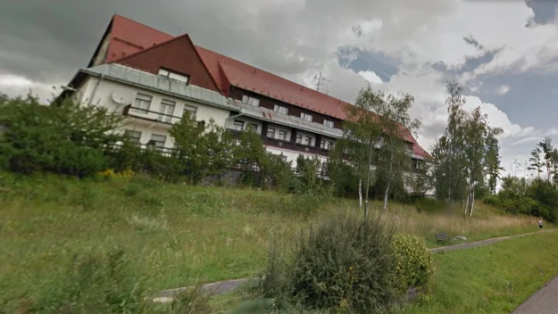 Duży budynek o czerwonym dachu otoczony drzewami oraz bujną zielenią, na tle pochmurnego nieba, w głębi widoczna ścieżka biegnąca wzdłuż trawnika.