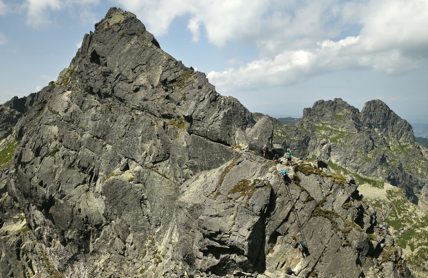 Niektóre odcinki na Orlej Perci są szczególnie niebezpieczne. Wymagają korzystania z łańcuchów i długich drabinek Strome szczyty górskie z wyraźnie widocznymi, surowymi skałami. Kilku turystów lub wspinaczy pokonuje skalistą trasę w słoneczne popołudnie, otoczeni majestatycznym krajobrazem.
