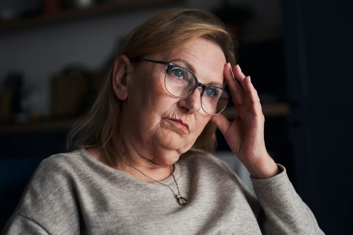 Starsza kobieta z okularami, siedząca zamyślona, podpiera głowę dłonią, nosi jasny sweter, na jej twarzy widoczny jest zamyślony lub zatroskany wyraz twarzy, tło jest rozmyte.