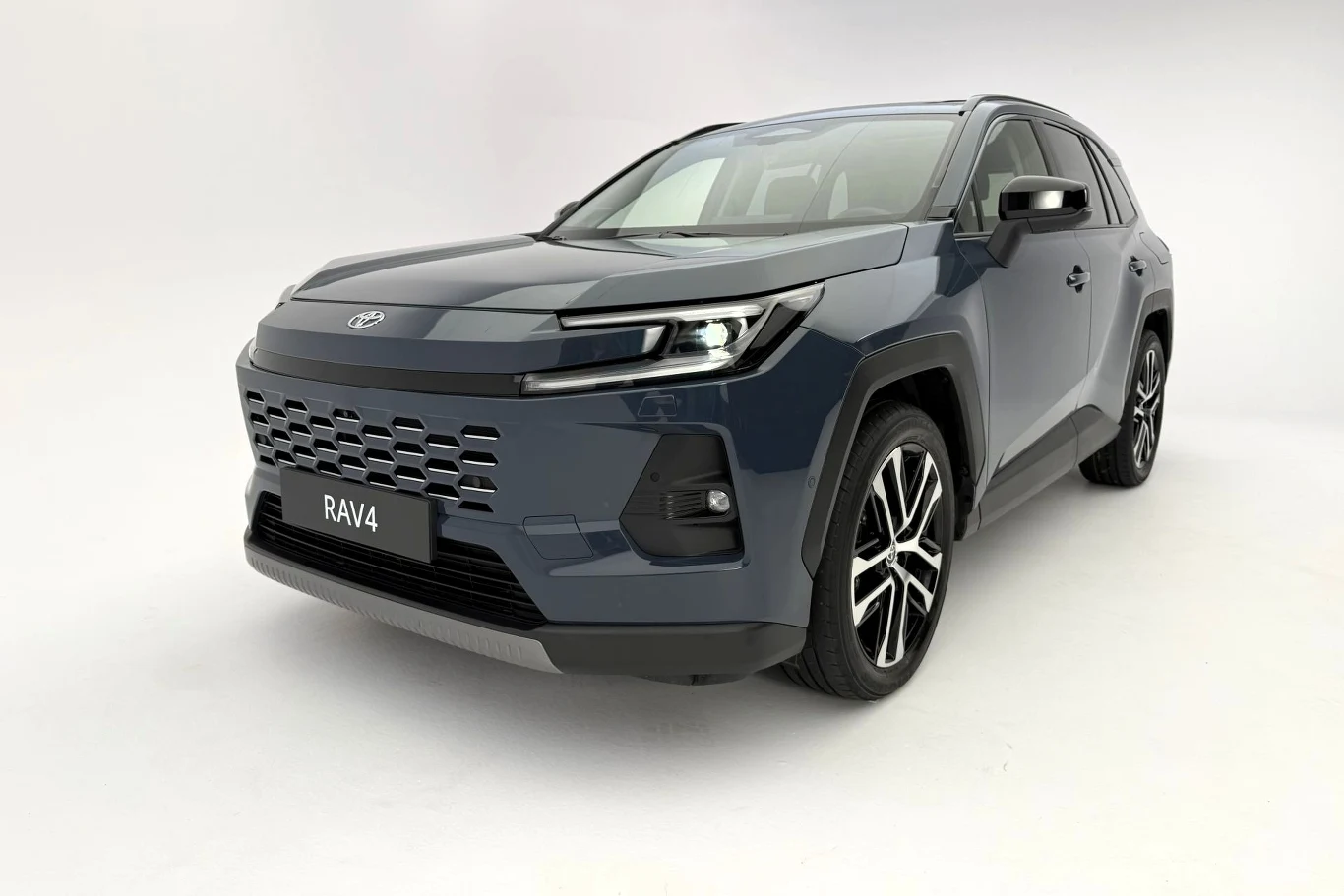 Nowa Toyota RAV4 wygląda świetnie i ma nowe układy hybrydowe. Szary SUV marki Toyota RAV4 o nowoczesnym designie na jednolitym, jasnym tle. Wyraźnie widoczna linia świateł LED, masywna krata chłodnicy oraz eleganckie felgi aluminiowe. Samochód jest zaparkowany, widok pod kątem z przodu.