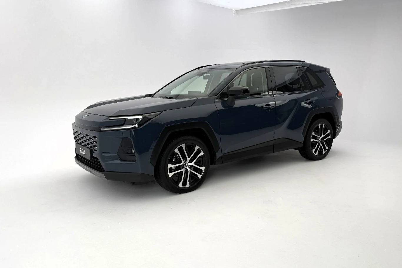 Nowa Toyota RAV4 zbudowana jest na tej samej, choć wzmocnionej, platformie, co poprzednia generacja. W efekcie samochód ma te same rozmiary: 4,6 m długości i 2,69 m rozstawu osi. Nowoczesny, niebieski SUV o dynamicznych liniach i dużych felgach, ustawiony na jasnym tle studyjnym, oba przednie światła LED włączone, pojazd wyeksponowany z lewej strony.