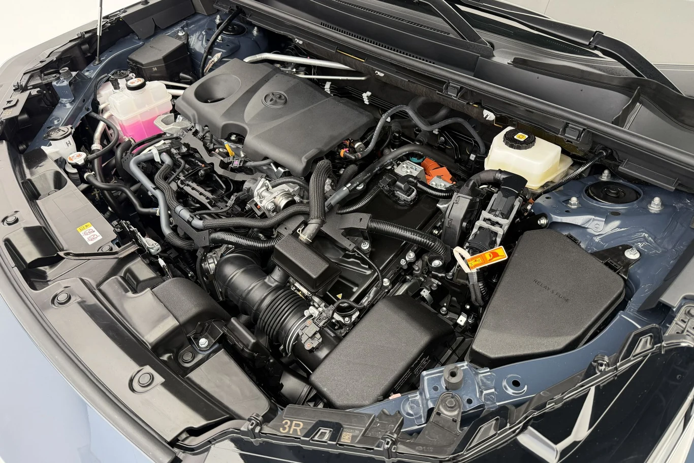Nowa Toyota RAV4 obecnie jest dostępna w Polsce tylko z układem hybrydowym. Do wyboru są dwa warianty: 183 KM z napędem na przód oraz 192 KM z napędem na cztery koła. Silnik samochodowy pod maską, widoczne różne elementy mechaniczne, przewody, zbiorniki płynów oraz pokrywa silnika z logo producenta.
