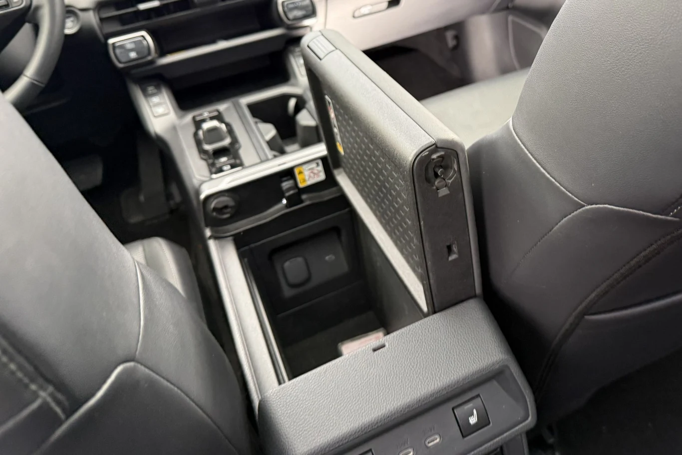 Pokrywa schowka między przednimi fotelami może otwierać się zarówno od strony kierowcy, jak i pasażera. Dodatkowo pokrywę można zdemontować, odwrócić i zamontować jako półkę na drobiazgi. Rozłożony środkowy podłokietnik z otwartym schowkiem w konsoli środkowej samochodu, widoczne detale wnętrza takie jak fotele, przyciski sterujące oraz uchwyty na napoje.