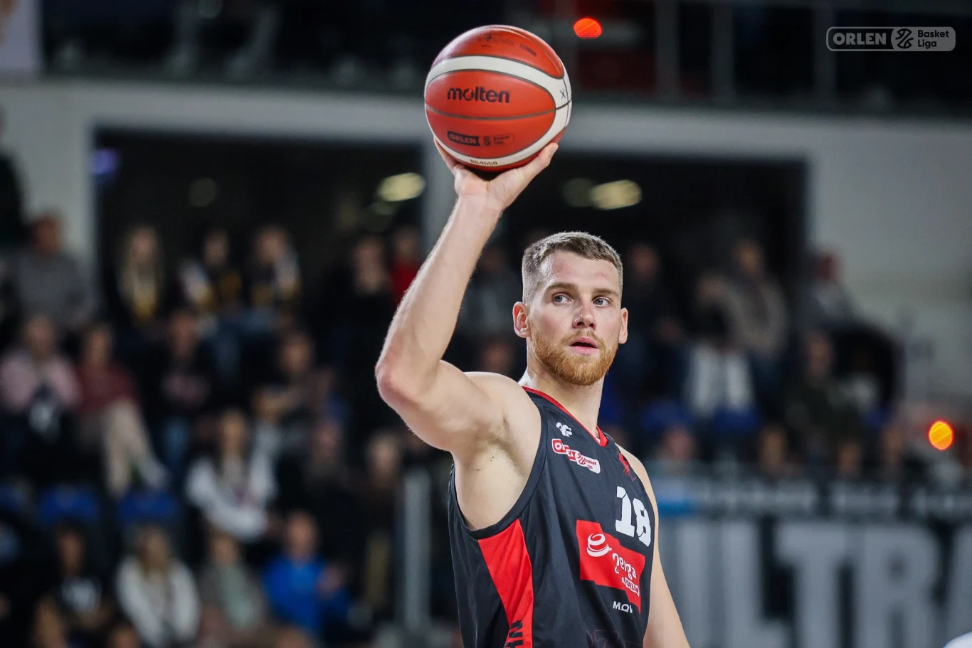 Koszykarz w czarno-czerwonym stroju z piłką uniesioną nad głową podczas meczu, w tle widownia zapełniona kibicami.