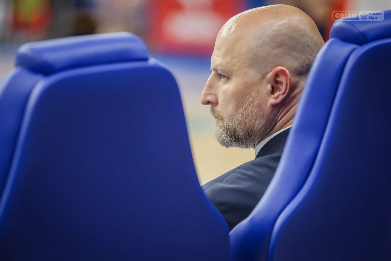 Łysa głowa mężczyzny z krótką brodą, ubranego elegancko w garnitur, widoczna zza dwóch niebieskich foteli, twarz skierowana w bok z poważnym wyrazem.