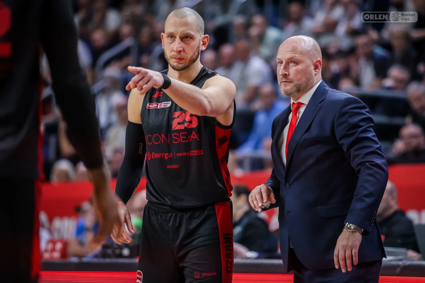 Zawodnik koszykarski w czarnym stroju drużyny z numerem 25 gestykuluje, wskazując ręką, obok niego stoi trener w garniturze z czerwoną krawatą. W tle widoczny tłum kibiców i fragment boiska.
