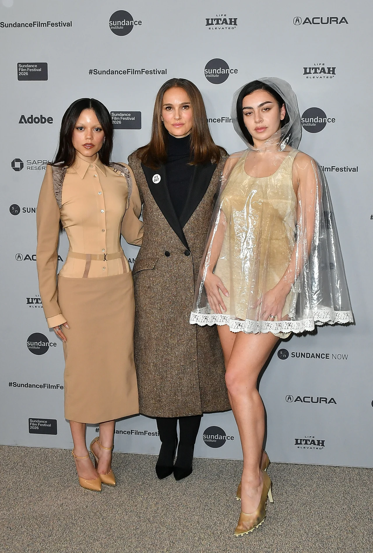 Trzy kobiety stoją na tle ścianki z logotypami partnerów festiwalu, ubrane w oryginalne kreacje. Jedna z nich ma na sobie transparentowy płaszcz nałożony na jasną sukienkę, druga beżowy kostium, a trzecia długi, elegancki płaszcz.