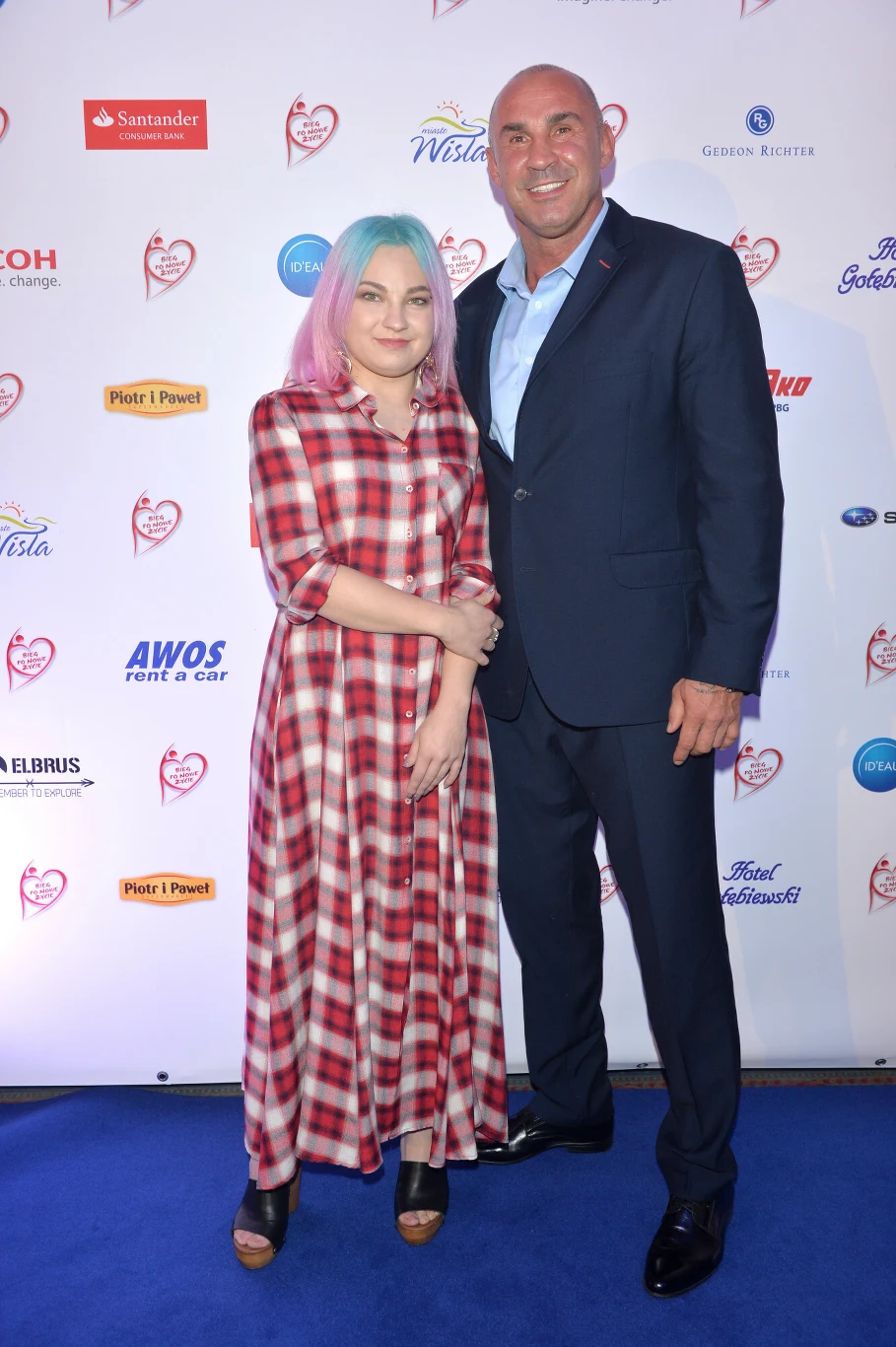 Dziewczyna o wielobarwnych włosach w czerwono-białej sukience w kratę stoi obok mężczyzny w ciemnym garniturze na tle ścianki z logotypami sponsorów i organizatorów wydarzenia.