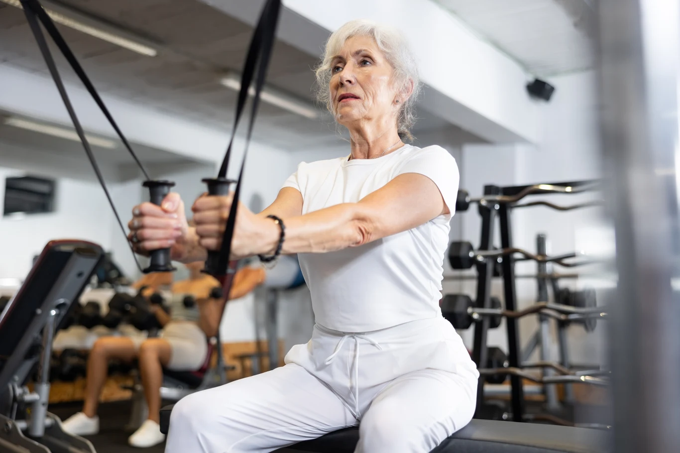 Starsza kobieta w białym stroju sportowym ćwiczy na maszynie do wyciskania w siłowni, koncentrując się na treningu. W tle widoczne są inne osoby i sprzęt fitness.