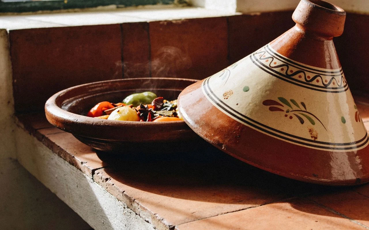 Tagine - tradycyjna marokańska potrawa Tradycyjny gliniany tagine częściowo przykryty ozdobną pokrywą, z widoczną parą unoszącą się nad soczystymi warzywami i mięsem w środku, ustawiony na kafelkowym blacie w naturalnym świetle.