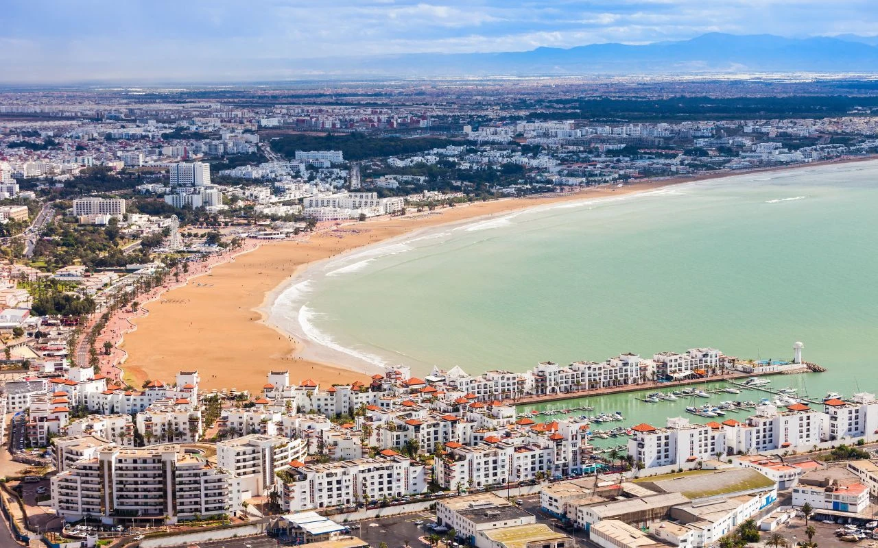 Agadir Panorama miasta z szeroką, piaszczystą plażą, zatoką, nowoczesnymi budynkami, mariną jachtową i rozciągającą się w tle zabudową miejską oraz górami na horyzoncie.