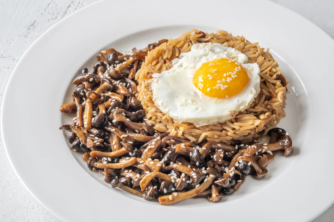 Grzybki shimeji świetnie komponują się z różnymi potrawami. Wzbogacają smak ryżu, makaronów i mięs Okrągłe gniazdo z makaronu orzo, w środku którego znajduje się sadzone jajko, obok duszone grzyby posypane białym sezamem na białym talerzu.