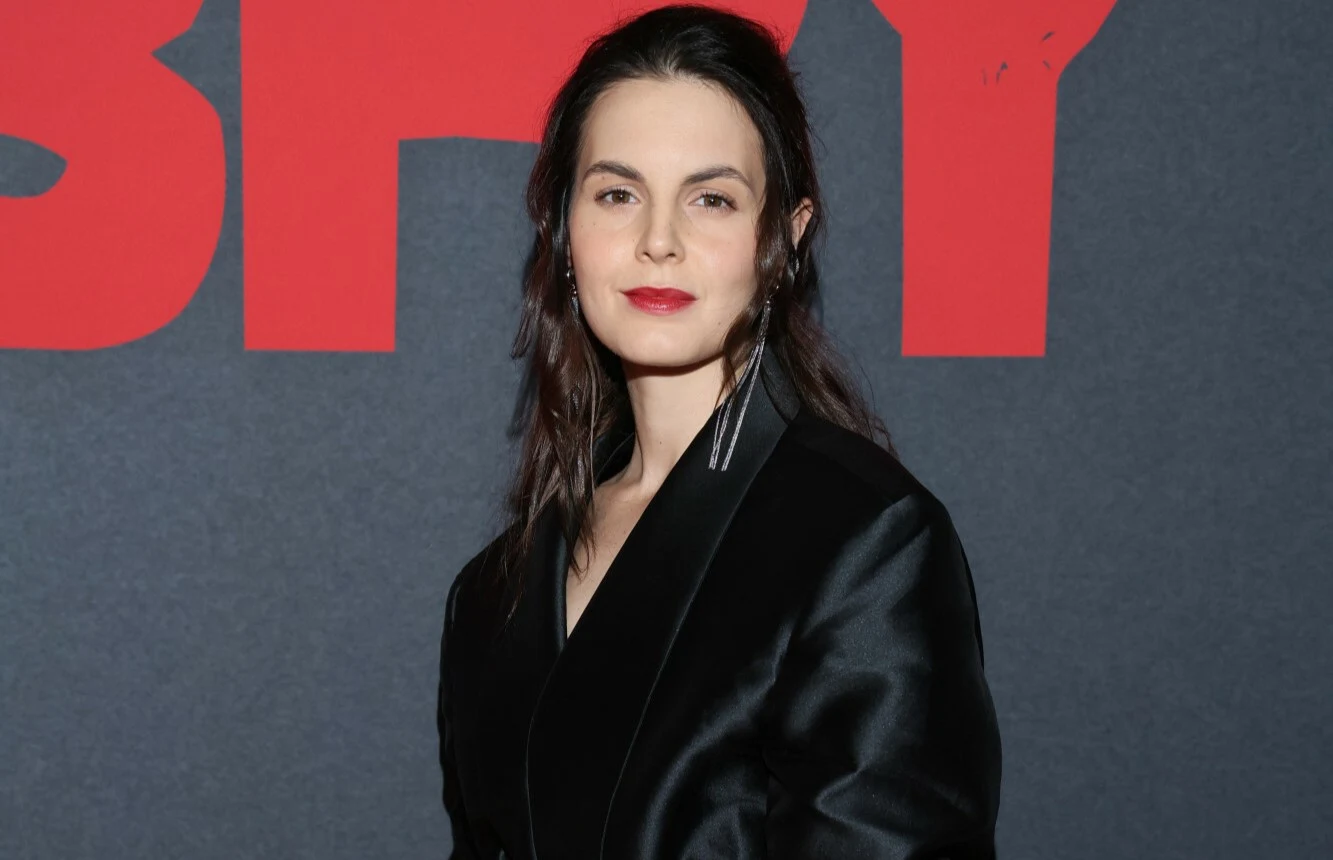 Agata Turkot Kobieta o ciemnych włosach w eleganckiej, czarnej marynarce stoi na tle ciemnej ścianki z dużym czerwonym napisem.