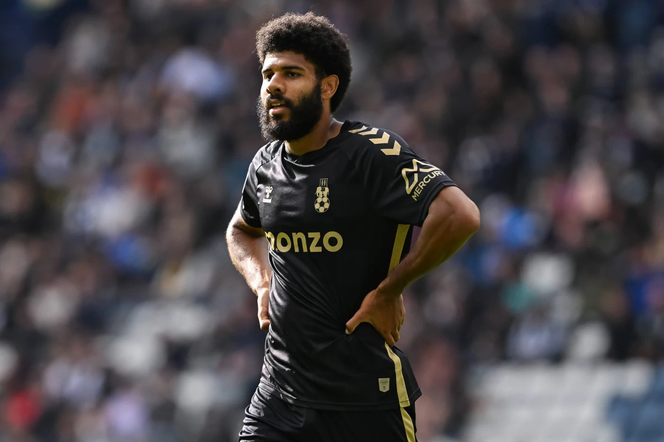 Piłkarz w czarnym stroju sportowym podczas meczu, stoi na boisku z rękami na biodrach, w tle rozmyta widownia stadionu.