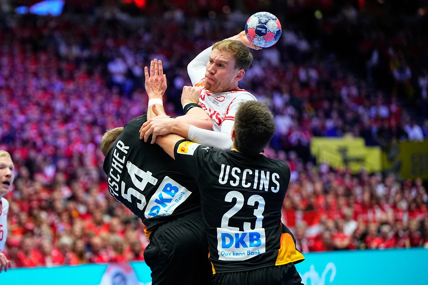 Dynamiczna akcja podczas meczu piłki ręcznej, zawodnik w białym stroju próbuje oddać rzut, będąc blokowanym przez dwóch obrońców w czarnych koszulkach. Na trybunach tłum kibiców w czerwonych barwach.