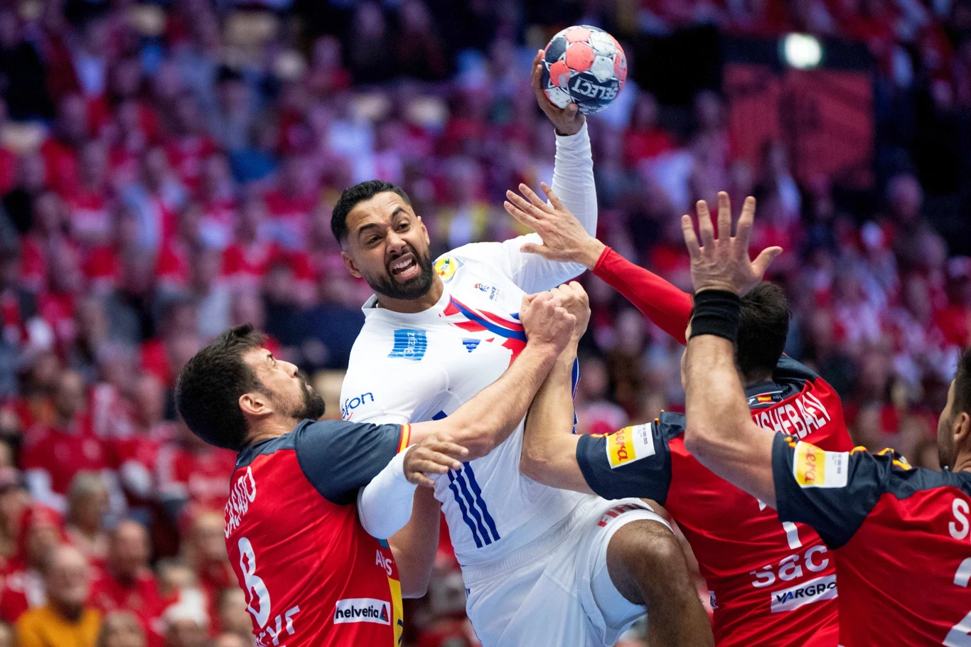 Zawodnik w białym stroju próbuje oddać rzut piłką do bramki, będąc blokowanym przez dwóch rywali ubranych w czerwone koszulki na boisku sportowym, wokół tłum kibiców.