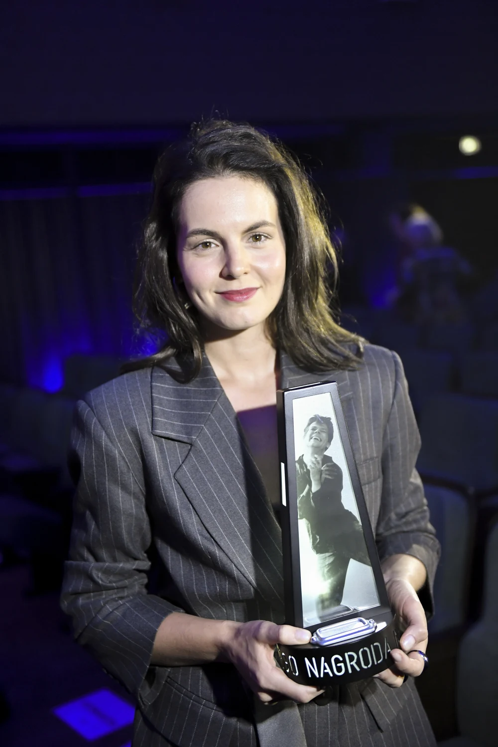 Agata Turkot Kobieta w eleganckiej marynarce trzyma w dłoniach nagrodę z podobizną postaci na plakietce, stojąc w oświetlonym miejscu na tle ciemnej sali.
