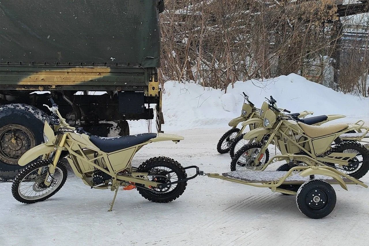 Iż-Enduro waży około 100 kilogramów, a jego bateria litowo-jonowa pozwala przejechać do 100 km Wojskowe motocykle terenowe w beżowym kolorze stoją na zaśnieżonym terenie, jeden z nich wyposażony w przyczepkę. W tle widoczny jest wojskowy pojazd ciężarowy oraz kolejne motocykle ustawione obok siebie, całość otoczona zimową scenerią.
