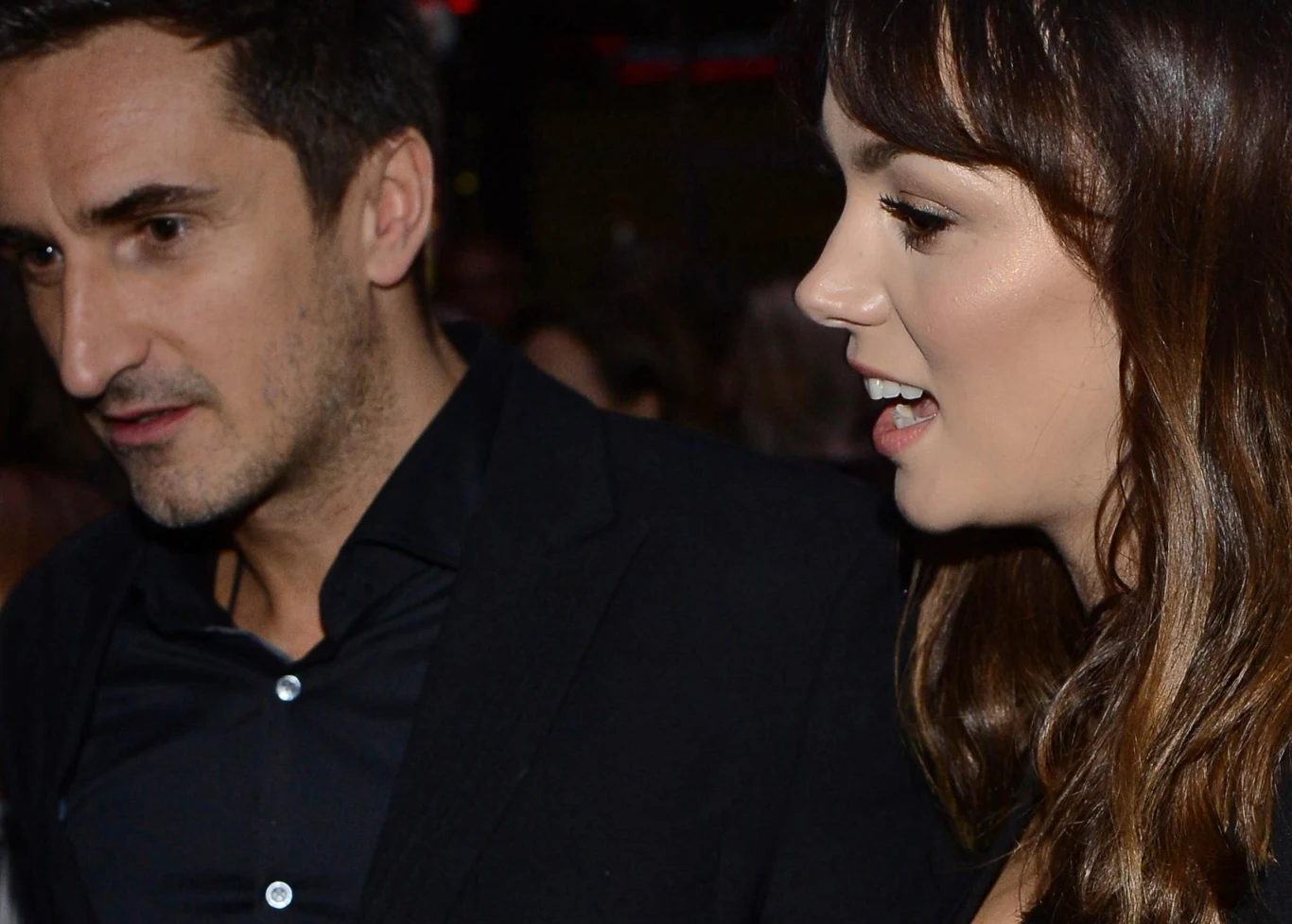 Sebastian Karpiel-Bułecka, Paulina Krupińska Para elegancko ubranych osób w półmroku, kobieta o brązowych włosach uśmiecha się i mówi, a mężczyzna o ciemnych włosach patrzy w bok, oboje znajdują się w tłumie ludzi.