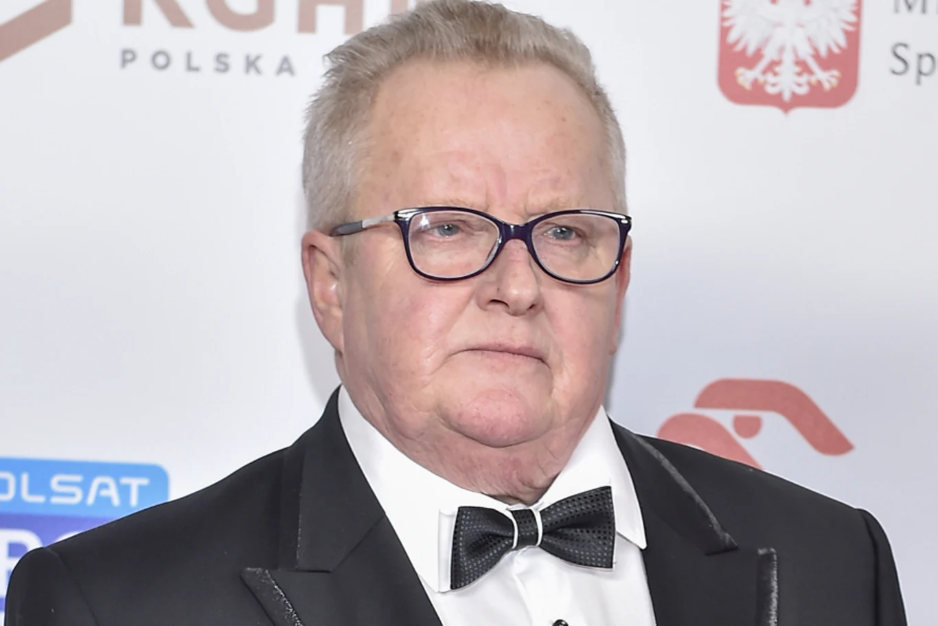 Mężczyzna w okularach i eleganckim smokingu z muchą pozuje na tle ściany z logotypami i godłem Polski.