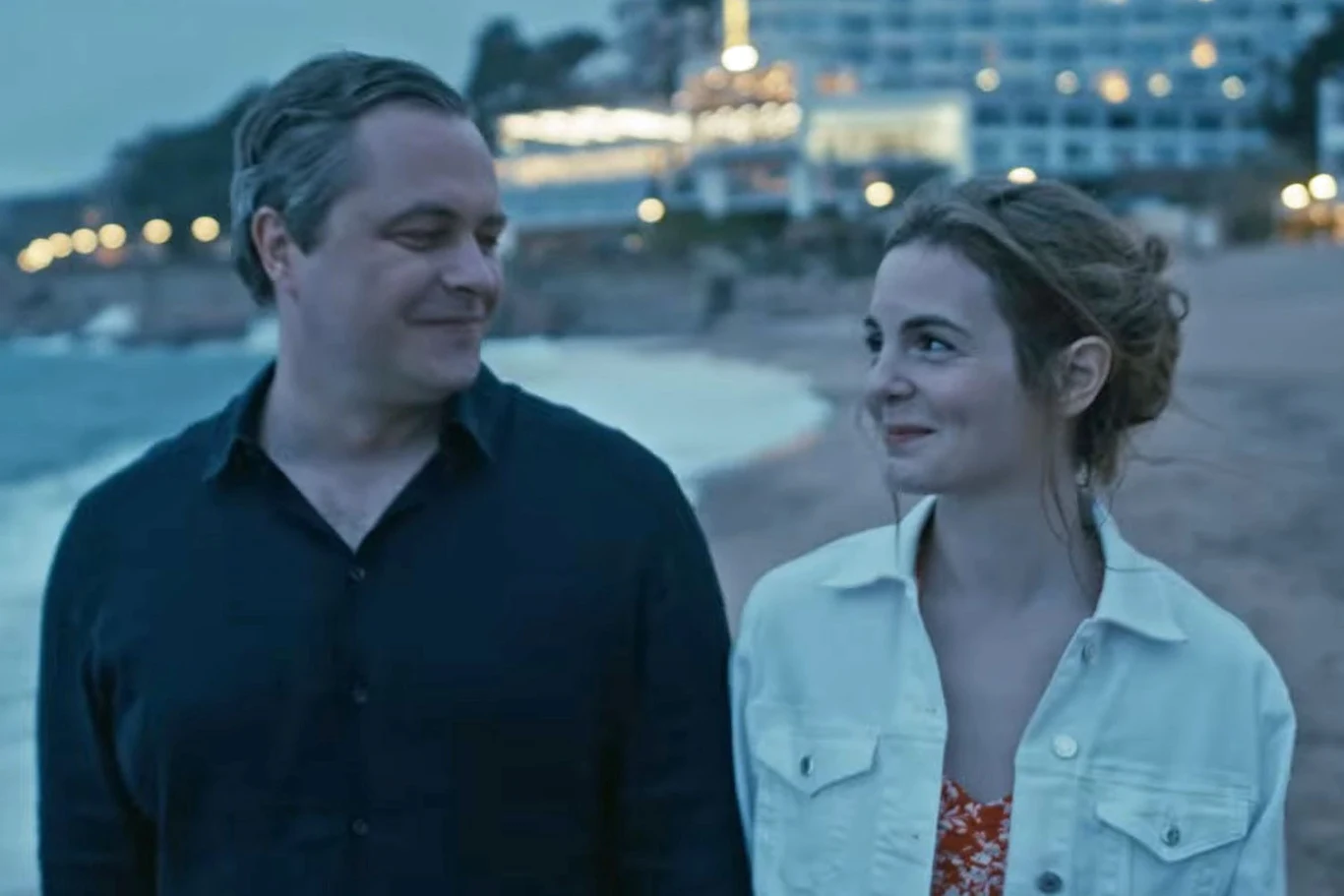 Para dorosłych spaceruje brzegiem plaży wieczorową porą, patrząc na siebie z uśmiechem. W tle widać rozświetlony budynek oraz delikatne światła na wzgórzu.