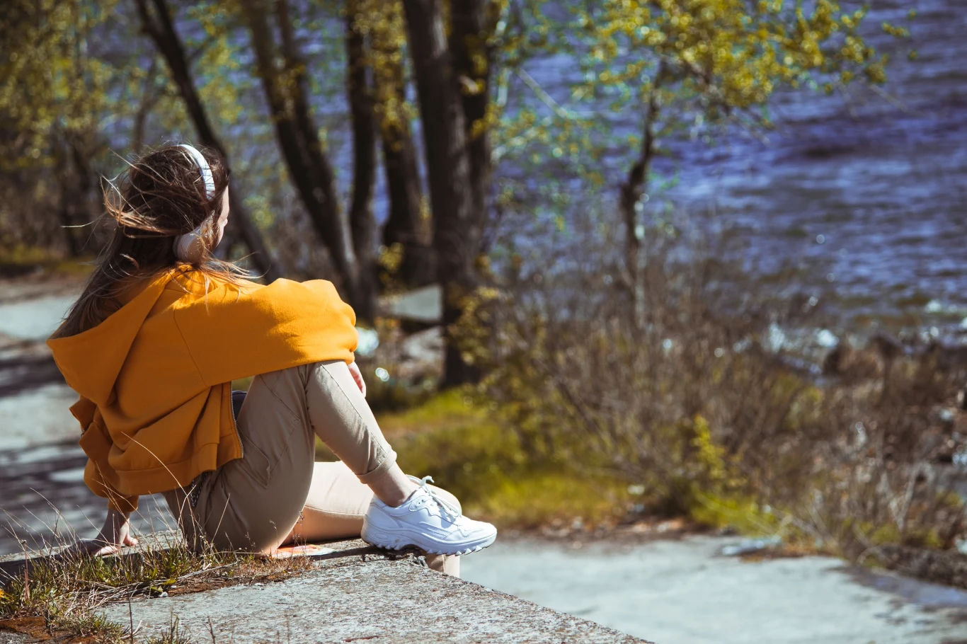 Dziewczyna w pomarańczowej bluzie i białych butach siedzi na betonowym brzegu, zwrócona w stronę rzeki lub jeziora, z widokiem na drzewa i wodę w tle. Cicha, spokojna atmosfera sprzyja refleksji.
