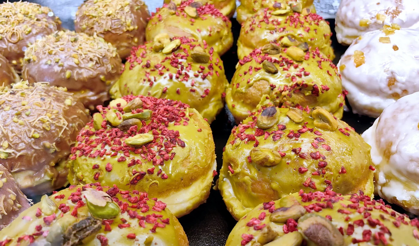 Kolorowe pączki z lukrem, posypane kawałkami pistacji oraz suszonych malin, ułożone w kilku rzędach na blacie.