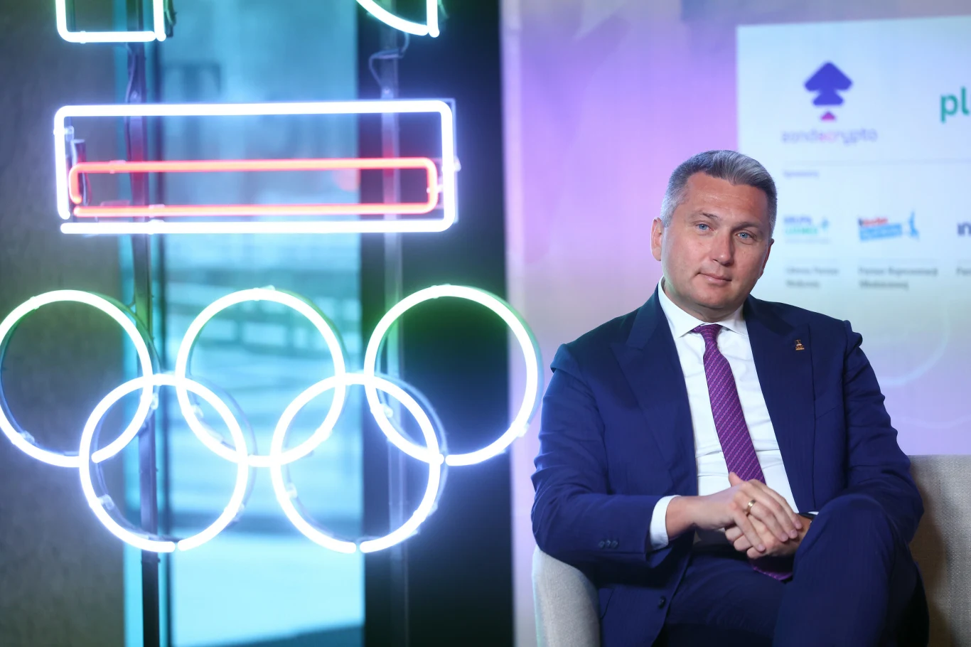 Radosław Piesiewicz, prezes Polskiego Komitetu Olimpijskiego Mężczyzna w garniturze siedzący na krześle obok świecącego neonowego symbolu kół olimpijskich na tle tablicy z logotypami partnerów i sponsorów.